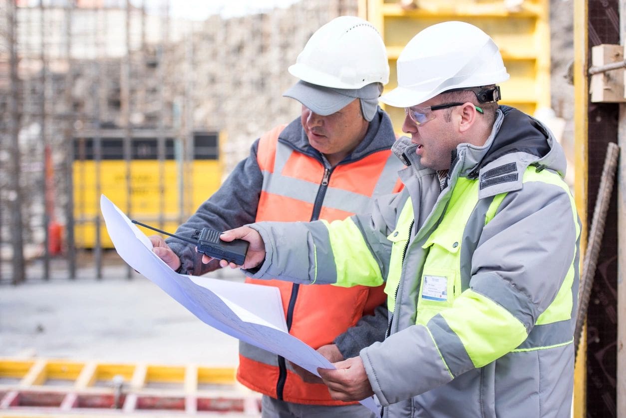 Two men in hard hats and safety vests looking at a paper.