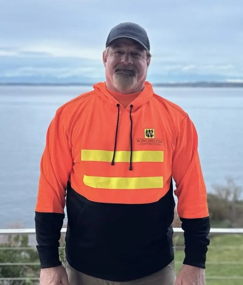 A man in an orange and black jacket standing next to the ocean.