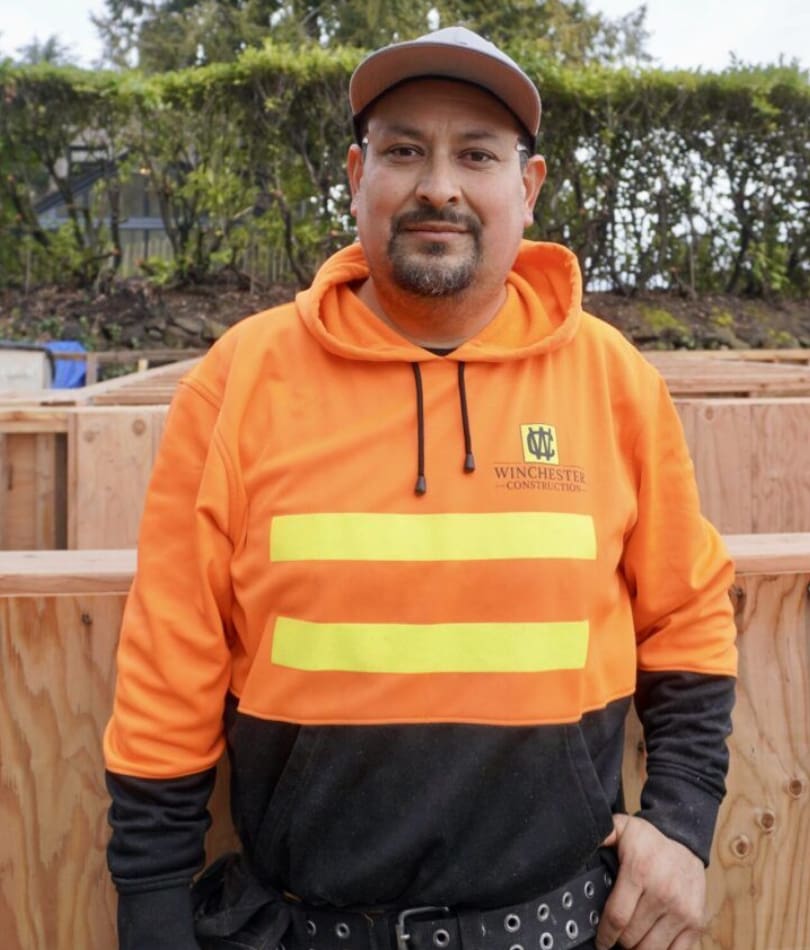 A man in an orange and black jacket standing next to a wooden fence.