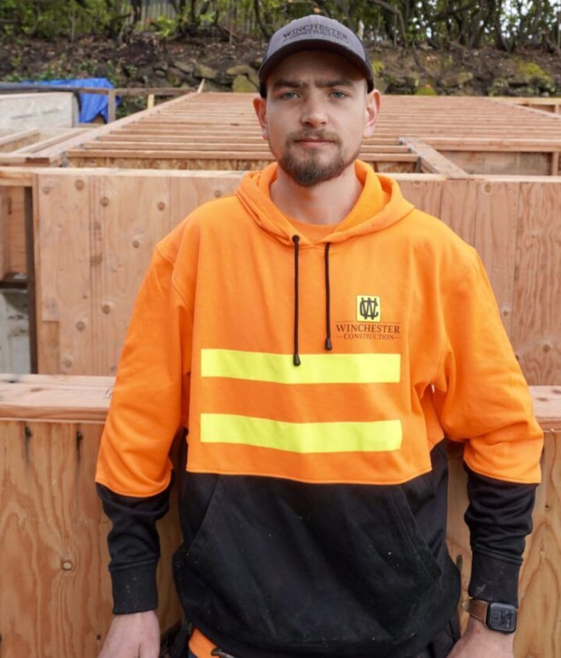 A man in an orange and black jacket standing next to wooden posts.